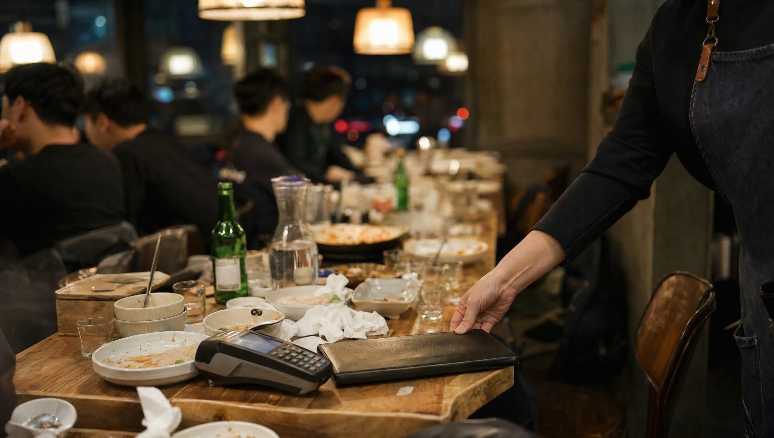 식당 안쪽 긴 테이블에 단체 손님 식사가 끝나고 계산서 폴더와 카드 단말기가 놓여 있는 장면